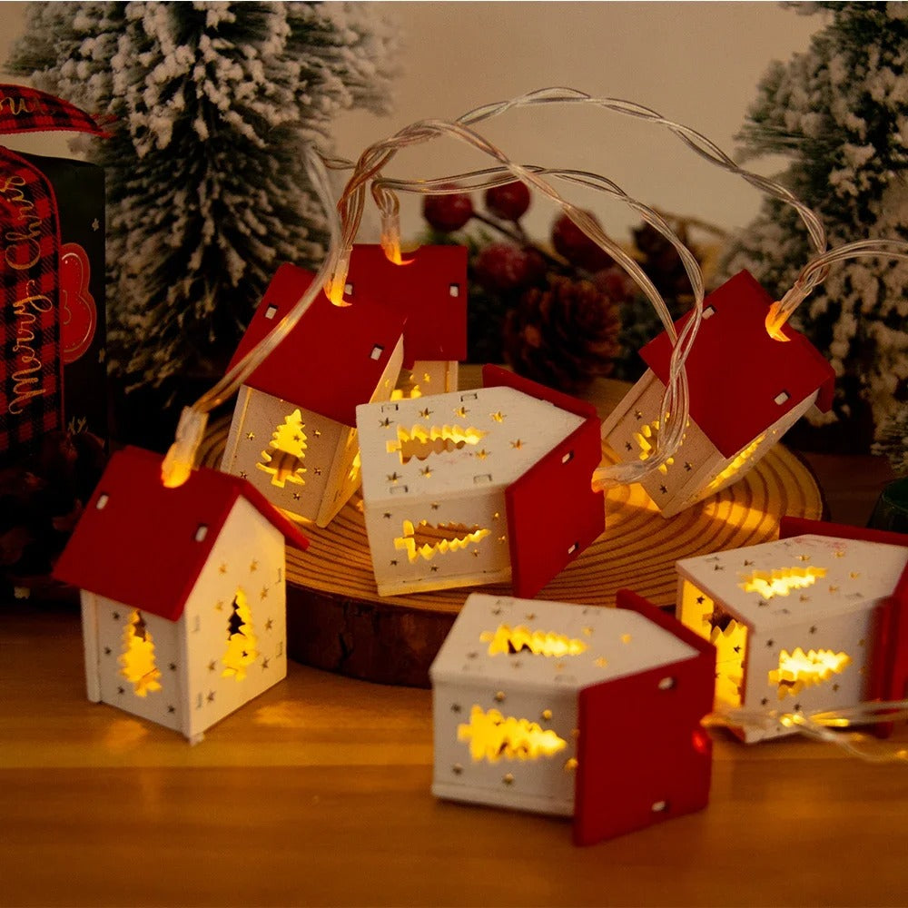 Decorative string lights shaped like small houses on a wooden surface with a Christmas tree in the background.