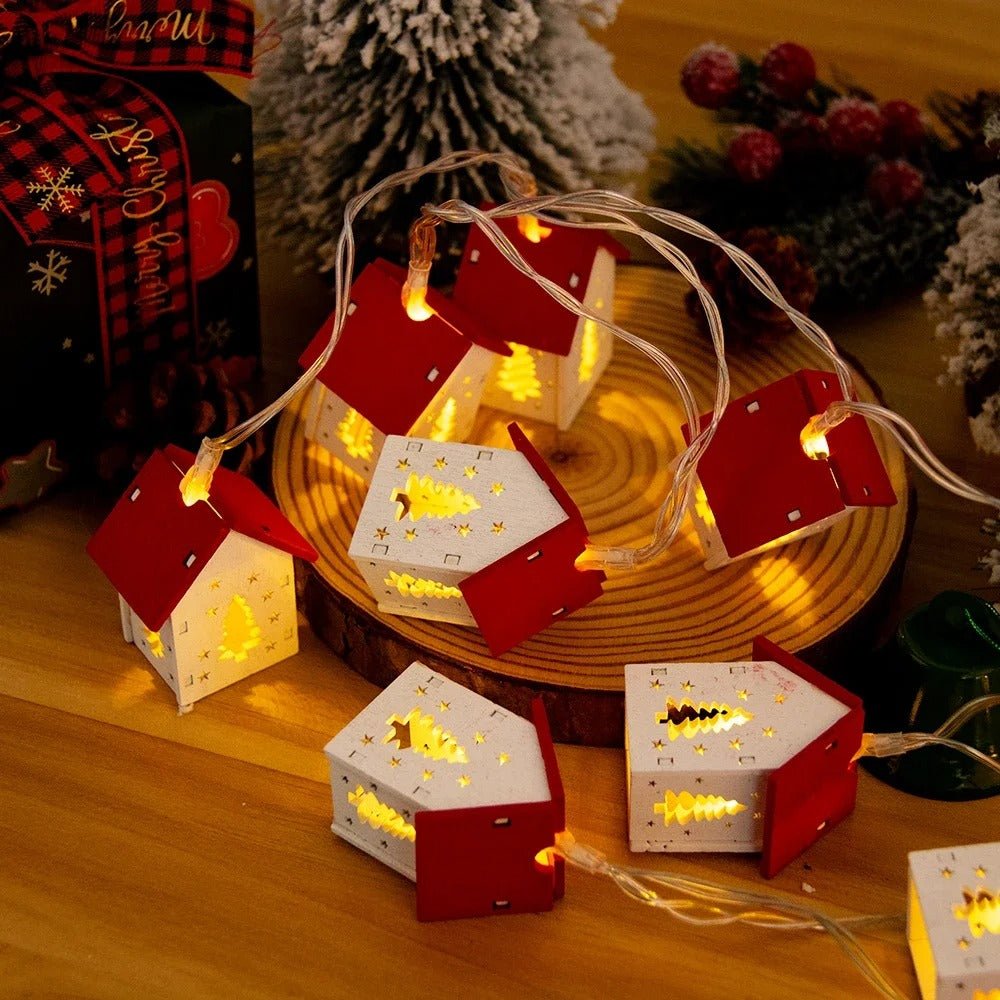 Decorative string lights shaped like small houses on a wooden surface with Christmas-themed decorations.