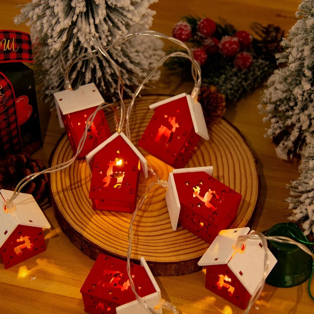 String lights shaped like red houses with white roofs on a wooden surface, surrounded by Christmas decorations.